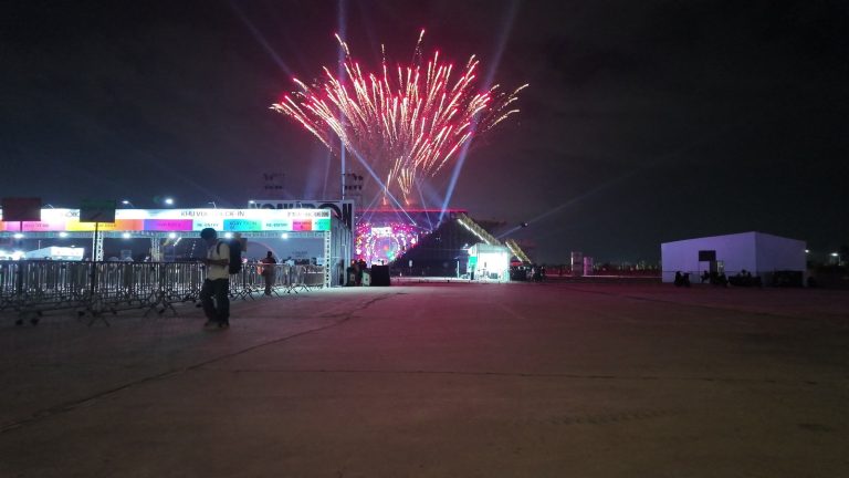 Pháo hoa bung sáng phía trên sân khấu concert ở Ocean City lúc đêm 6/9, các tia laser màu tím quét ngang trời, phía dưới là dãy cổng check-in nhiều màu