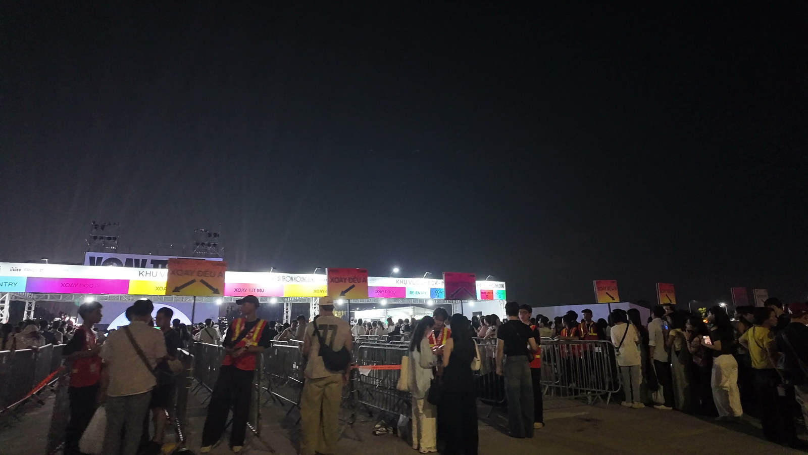 Đám đông khán giả trẻ xếp hàng dọc theo hàng rào kiểm soát trước cổng vào concert lúc trời đã tối, phía trên là dãy bảng hiệu nhiều màu của khu vực check-in