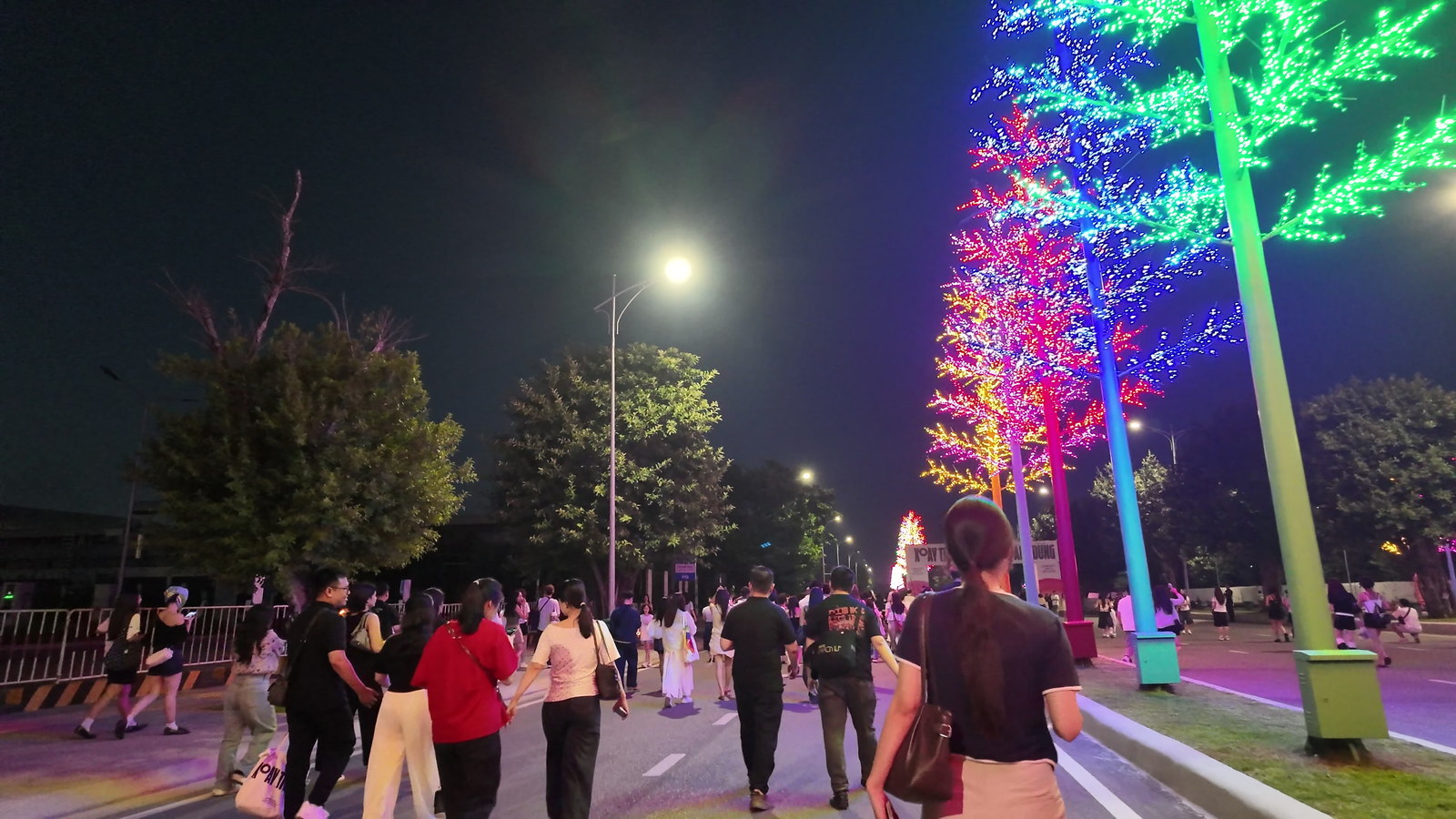 Người đi bộ trên trục đường lớn dẫn vào khu tổ chức concert buổi tối, hai bên là hàng cây trang trí đèn led nhiều màu, xa xa là ánh sáng của khu vực sân khấu