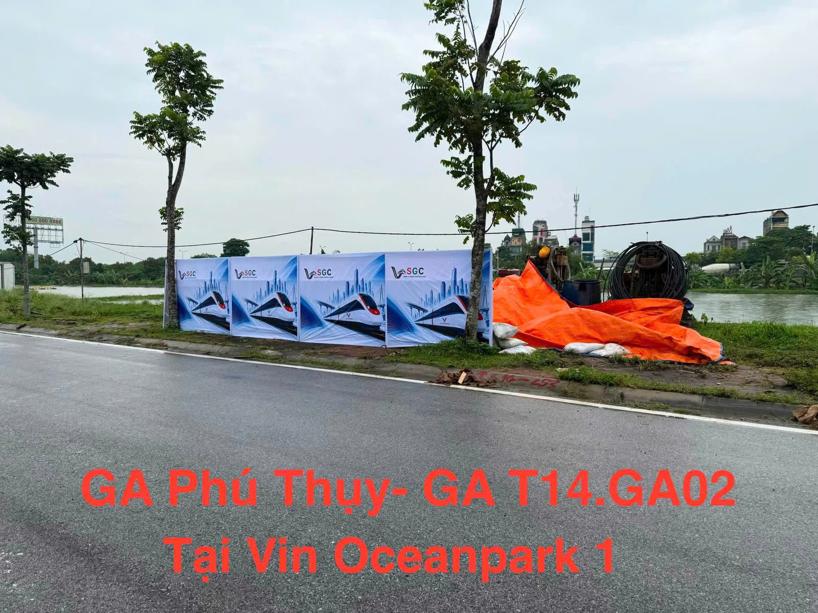 Hàng rào công trường nhà ga metro đặt ven đường tại khu vực Ocean Park 1, in phối cảnh đoàn tàu VinSpeed, phía sau là cánh đồng và máy khoan thăm dò
