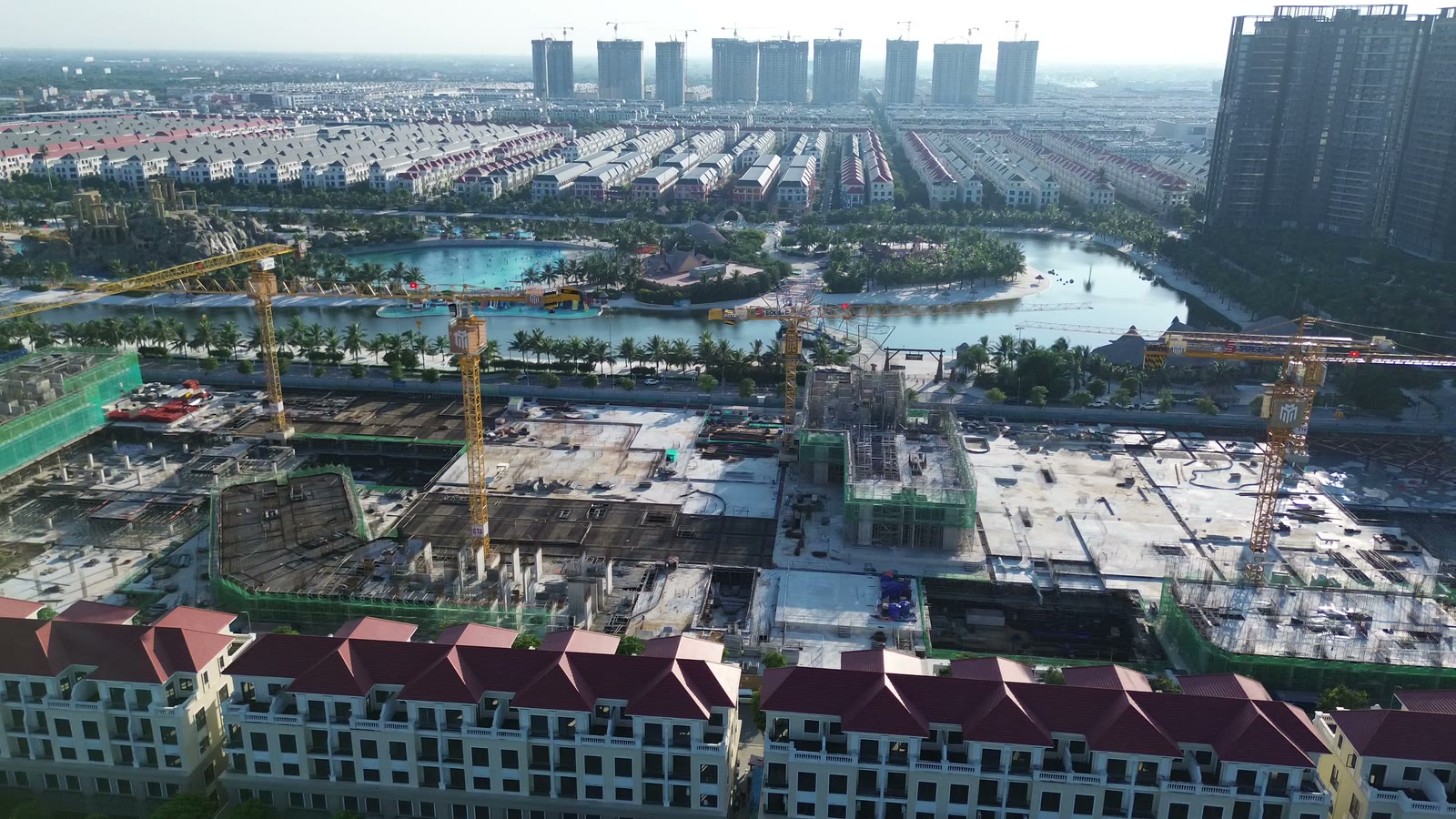 Công trường Masteri Grand Coast nhìn từ xa, phía sau là hồ tạo sóng và toàn cảnh khu đô thị Ocean Park với các dãy nhà thấp tầng và toà cao tầng