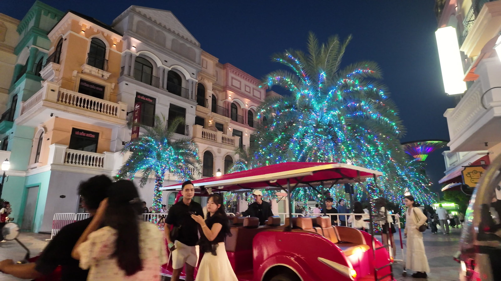 Dãy phố kiểu châu Âu ở Ocean City vào buổi tối, đèn vàng hắt lên các mặt tiền, một cây trang trí phát sáng xanh ở giữa, đông người đứng chụp ảnh và đi lại quanh khu vực