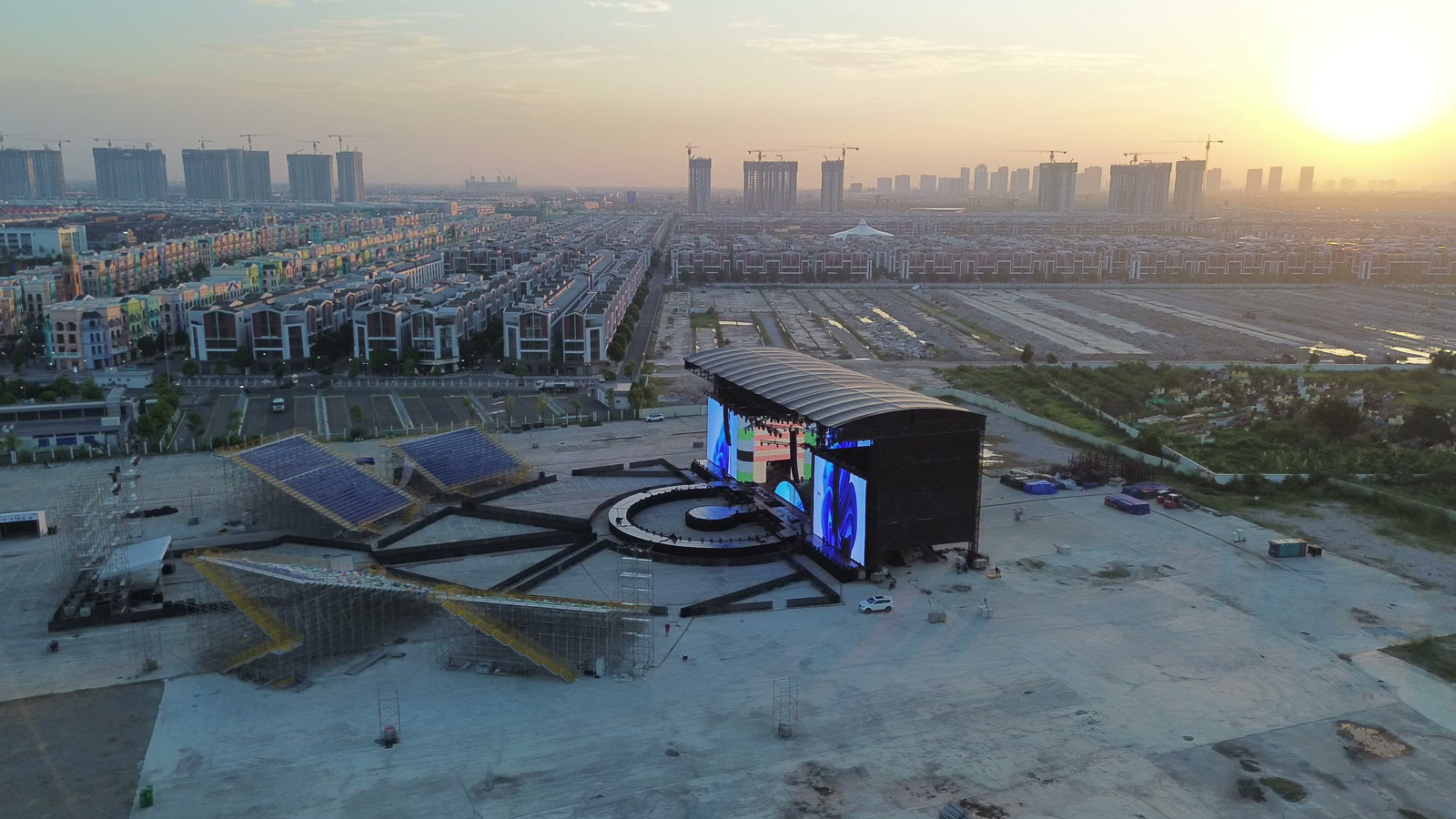 Toàn cảnh khu tổ chức concert lúc hoàng hôn nhìn từ flycam, sân khấu và khán đài nằm giữa mặt bằng rộng, bên trái là các dãy nhà thấp tầng san sát của khu đô thị, xa xa là những toà cao tầng đang xây