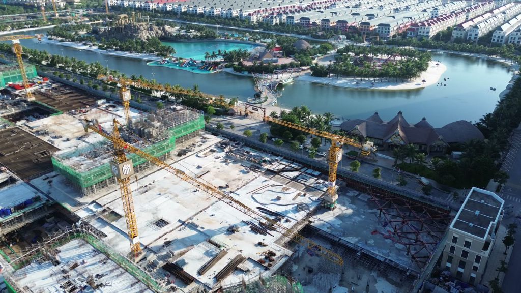Công trường Masteri Grand Coast nhìn từ flycam, các cần cẩu tháp Masterise Homes phía trước, hồ tạo sóng Wonder Wave Park với bãi cát và hàng dừa nằm ngay đối diện