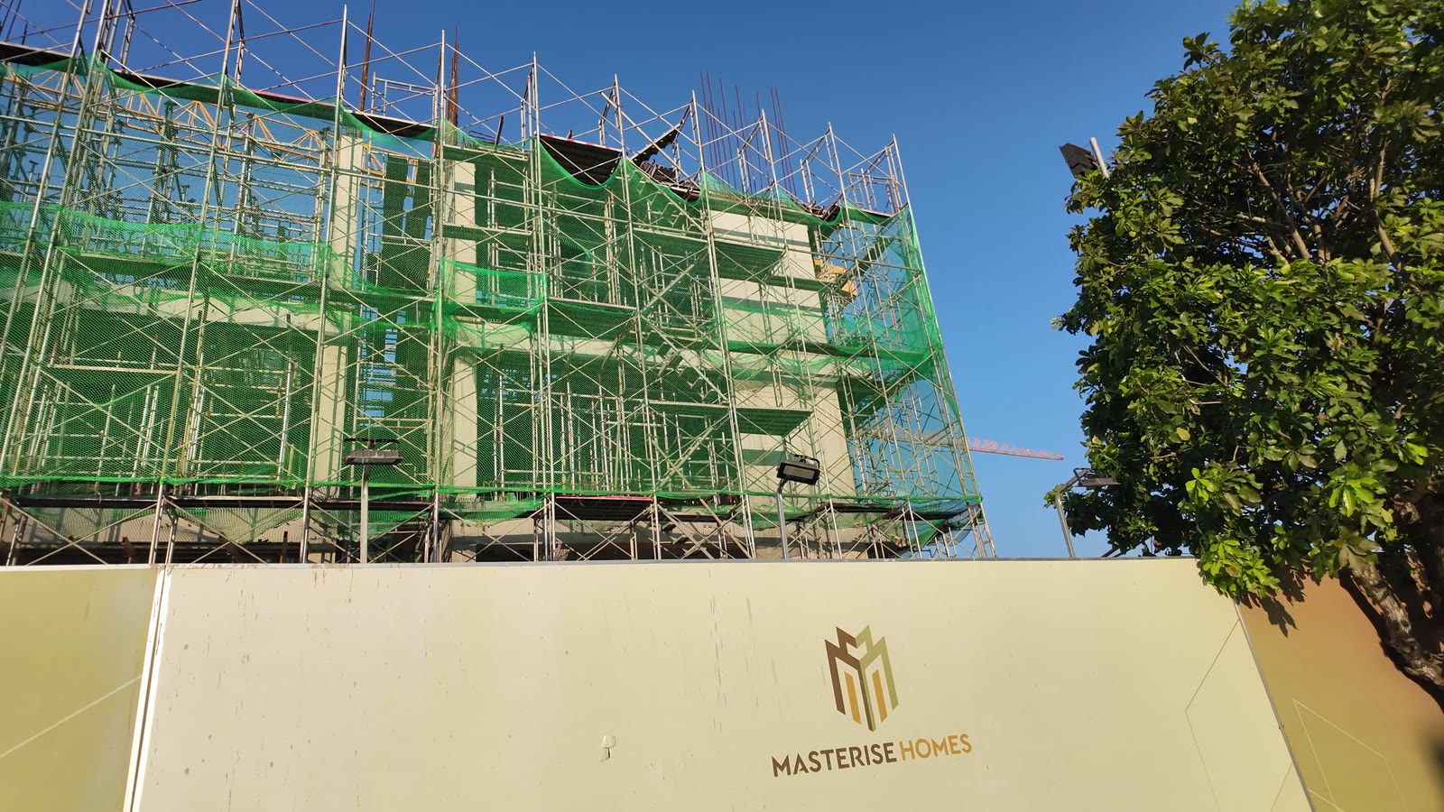 Nhìn từ dưới đường lên một toà căn hộ Masteri Grand Coast đang thi công, đếm được ba sàn bê tông sau lớp lưới an toàn màu xanh và giàn giáo, phía trước là hàng rào công trường in logo Masterise Homes