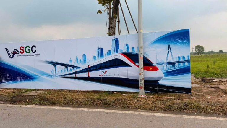 Hàng rào công trường nhà ga metro đặt ven đường khu vực Thanh Oai gần Sân vận động VinFast, in phối cảnh đoàn tàu VinSpeed, phía sau là cánh đồng